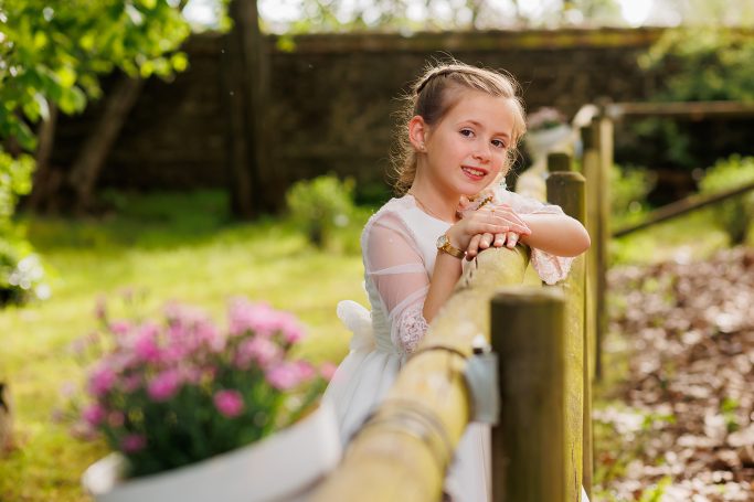 Niña en vestido blanco apoyada en una cerca, sonriendo, rodeada de naturaleza y flores.