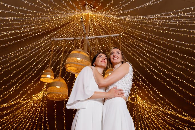 Dos mujeres en vestidos blancos abrazadas bajo luces decorativas brillantes.