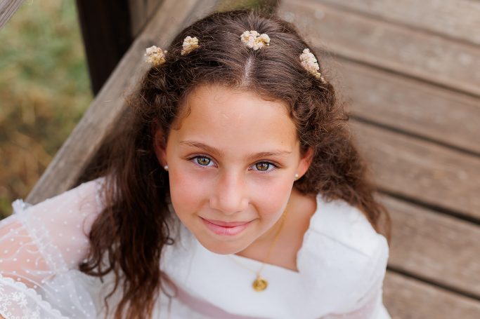 Niña con vestido blanco y flores en el cabello, sonriendo desde un puente de madera.