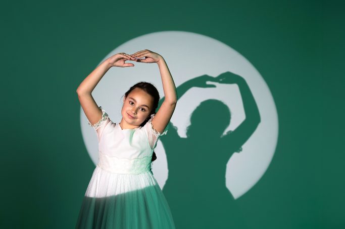 Niña en un vestido blanco formando un corazón con las manos, sombra proyectada detrás.